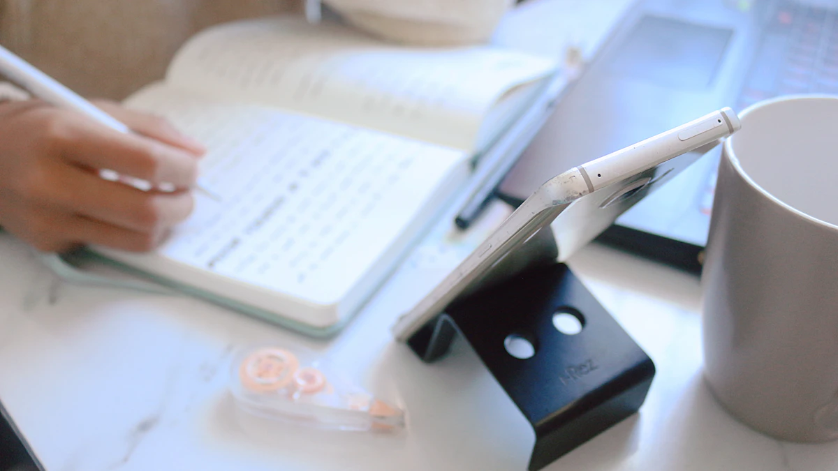Hand writing in a notebook beside a phone and mug on a white desk.
