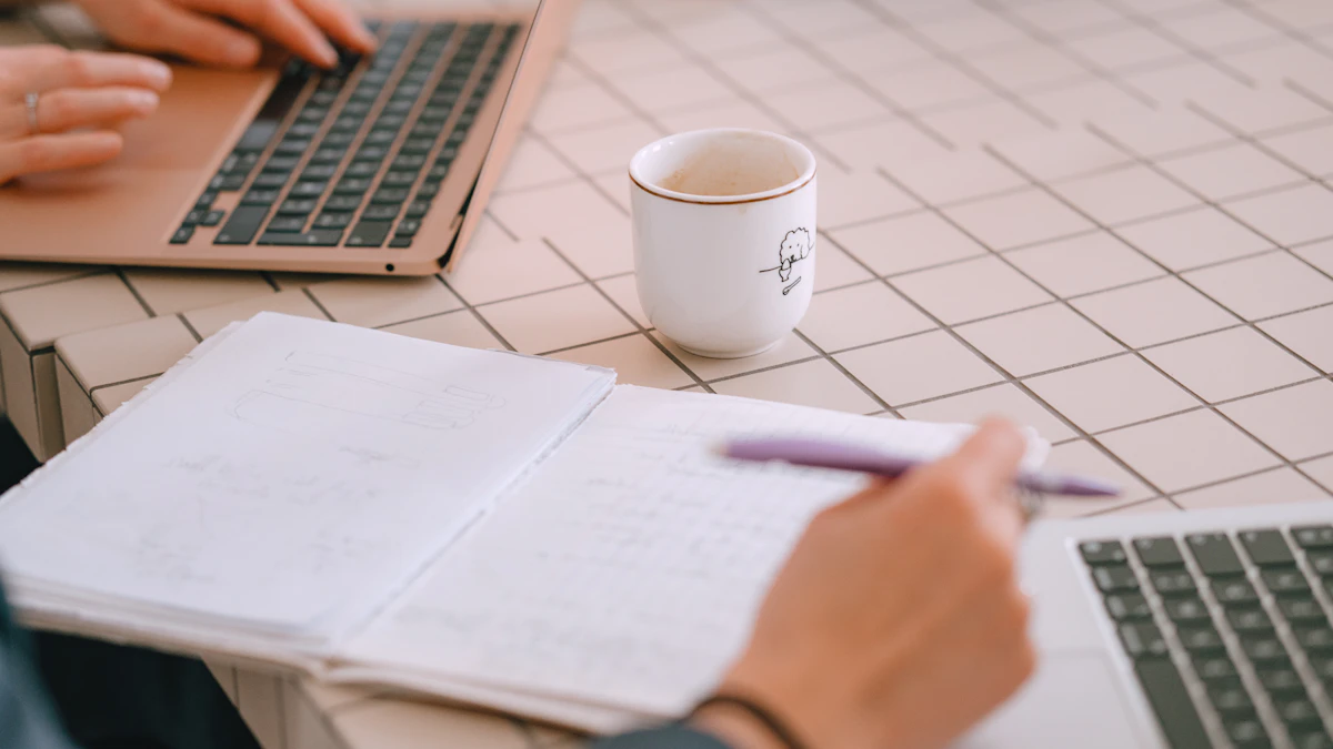 Two people working at a tiled table with laptops, an open notebook, a pen and a coffee mug.