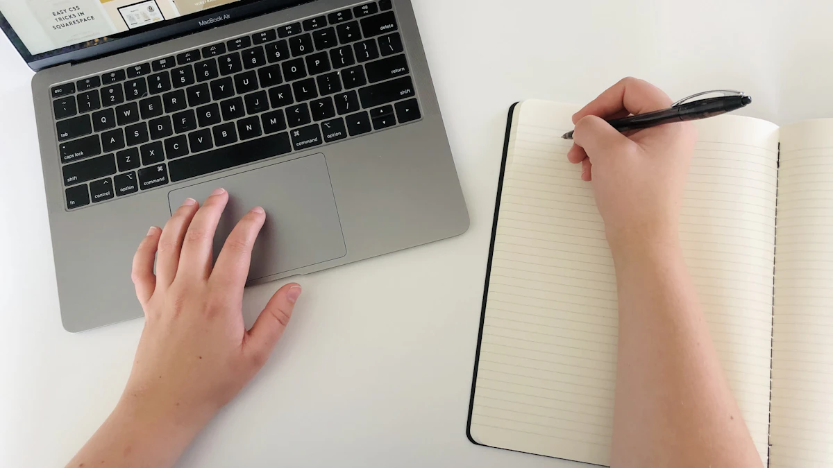 Overhead view of a person using a laptop and holding a pen over an open notebook.